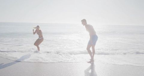 Couple jumping back from incoming waves on sunlit beach, backlit summer fun with reflections