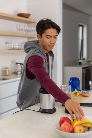 Asian man reaching for banana while assembling countertop blender on kitchen island