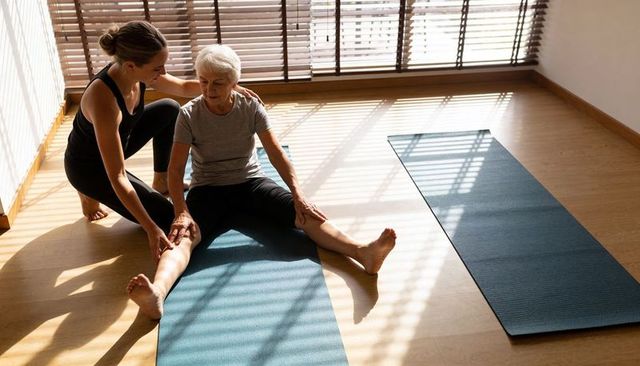 Young instructor helping senior woman stretch on yoga mat in sunlit studio for balance