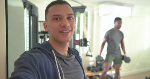 Diverse friends taking gym selfie while workout partner lifting dumbbells in home gym