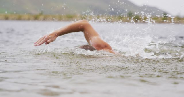 Triathlete swimming in a lake with mountains in the background, showing powerful arm stroke and creating splashes. Suitable for use in articles, advertisements, and promotional materials related to sports, endurance events, outdoor activities, and fitness training.