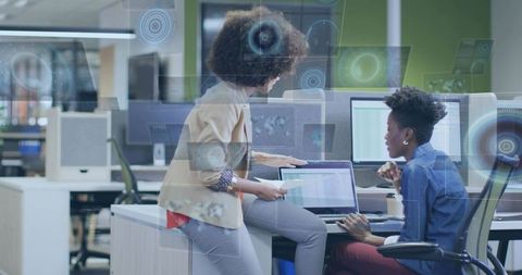 Female colleagues collaborating over laptop and analytics dashboard in open-plan office