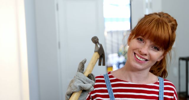 Young woman with red hair smiling and holding a hammer indoors, dressed in casual outfit with striped shirt and jeans overall. Perfect for use in content related to home improvement, renovation projects, DIY activities, team efforts in house construction, and promoting a can-do approach in domestic settings.