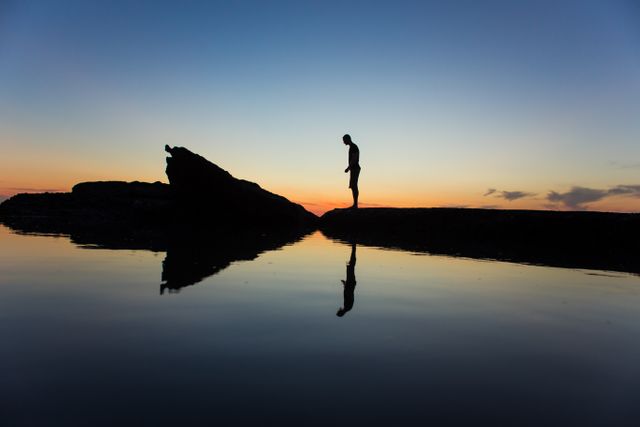 Silhouette of a man is reflected in smooth water while standing near dramatic rocks during sunrise. Tranquil and serene, ideal for concepts of solitude, reflection, nature's beauty, and beginnings. Suitable for inspirational posters, calm and peaceful themes, and mindful meditation visuals.