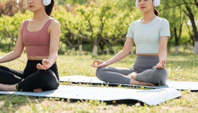 Two women meditating in park on yoga mats wearing headphones for outdoor mindfulness