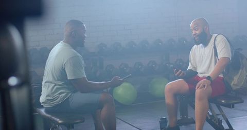 Two men chatting on gym benches during workout break holding smartphones near dumbbells
