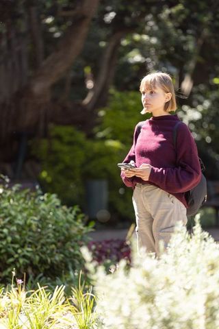 Young woman standing in sunlit park holding smartphone and backpack, thoughtful student