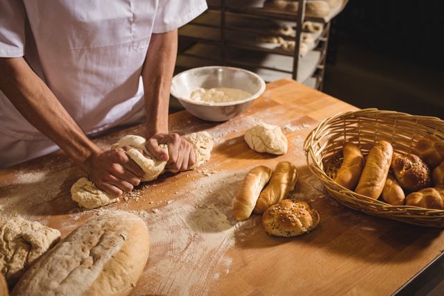 Baker kneading dough on wooden surface in artisan bakery. Freshly baked bread and rolls are visible. Ideal for use in culinary blogs, bakery advertisements, cooking tutorials, and food-related publications.