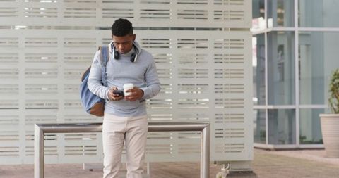 African american commuter standing at rail checking phone holding coffee and backpack