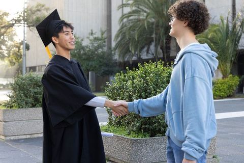 Sunlit asian graduate shaking hands with caucasian friend on campus celebrating achievement