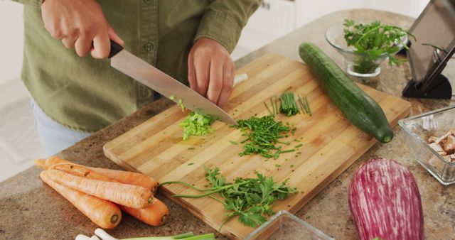 Person chopping fresh vegetables on a wooden cutting board in a kitchen. Visible ingredients include carrots, cucumber, herbs, and other fresh produce. This can be used for content related to cooking, recipes, healthy eating, food preparation, and kitchen activities.