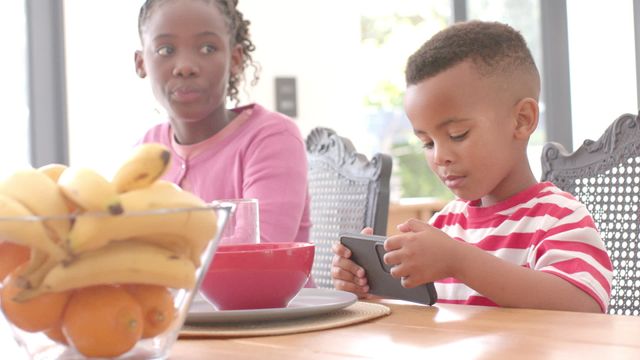 Young brother engrossed in using a smartphone while sister enjoys breakfast in the kitchen. Ideal for themes around family interactions, morning routines, modern technology use among children, and cozy kitchen settings.