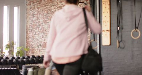 Woman entering industrial gym carrying duffel wearing pink hoodie near dumbbell rack
