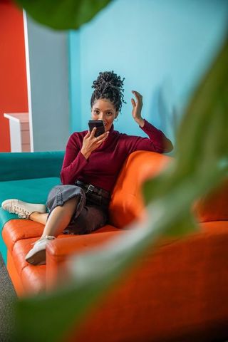 African american woman speaking into smartphone while relaxing on vibrant orange sofa