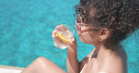 Sunlit woman sipping lemon water at turquoise poolside in clear sunglasses - resort glamour