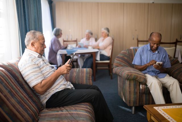 Senior men are sitting on couches using mobile phones in a nursing home common area. In the background, elderly women are having coffee and chatting at a table. This image can be used to depict the integration of technology in elderly care, social activities in retirement homes, and the modern lifestyle of seniors. It is suitable for articles, advertisements, and brochures related to senior living, elderly care, and technology for seniors.