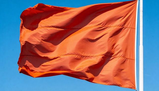 Vibrant orange flag fluttering against clear blue sky with stitched seams and white pole