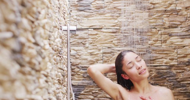 This image shows a woman enjoying a relaxing shower in a beautifully designed stone-walled bathroom. Water cascades down as she experiences a moment of peace and tranquility. Ideal for promoting spa treatments, wellness products, skincare, and self-care routines.