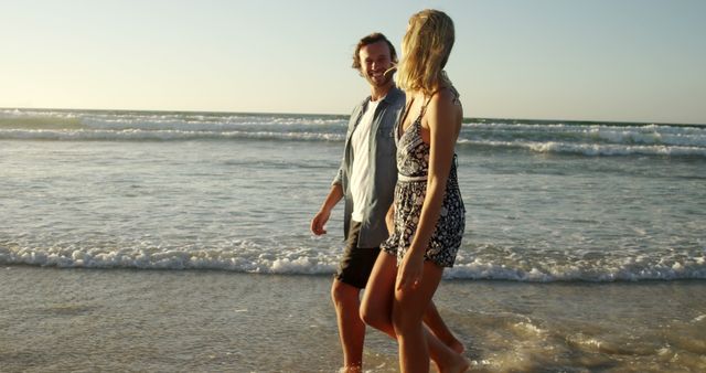 A young Caucasian couple enjoys a romantic walk along the beach at sunset, with copy space. Their casual attire and relaxed demeanor suggest a peaceful moment shared between them.