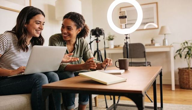 Female creators collaborating on content creation at home using laptop, ring light, camera