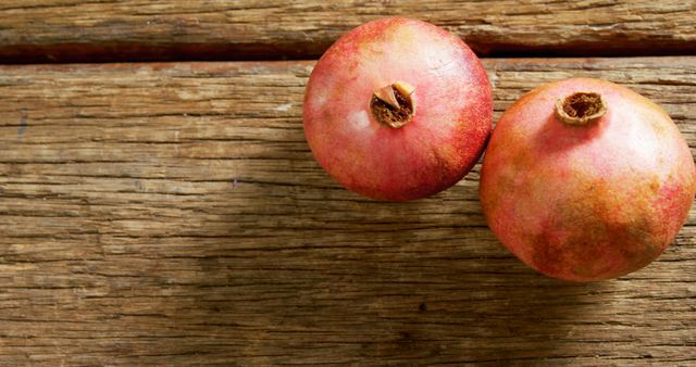 Two ripe pomegranates rest on a rustic wooden surface, showcasing their vibrant red skin. Their placement offers a natural and organic feel, highlighting the fruit's association with health and vitality.