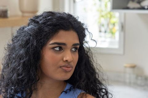 Indian woman gazing toward window in sunlit kitchen, curly hair serene close-up portrait