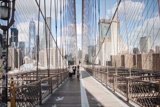 People walking on Brooklyn Bridge with the iconic New York City skyline in the background. Ideal for travel guides, tourism advertisements, cityscape photography, articles about New York, and urban architecture projects.