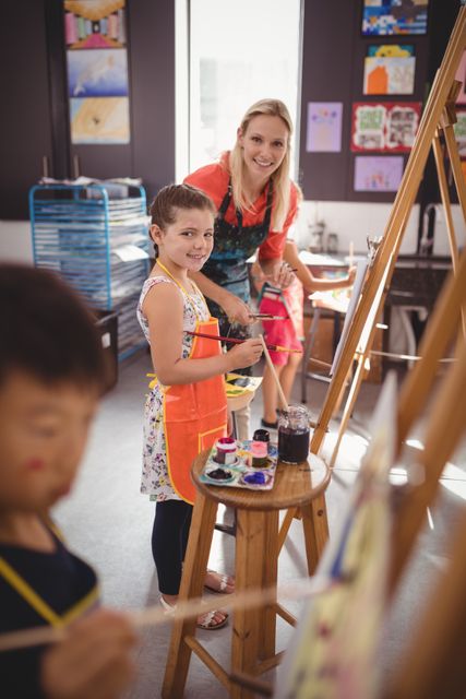 Teacher assisting young girl in art class at school. Girl wearing apron, painting on easel. Classroom filled with art supplies and children's artwork on walls. Ideal for educational content, school brochures, art class promotions, and creative learning materials.