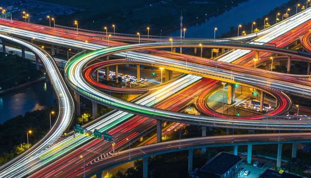 Night aerial highway interchange streaming neon light trails on curving elevated ramps