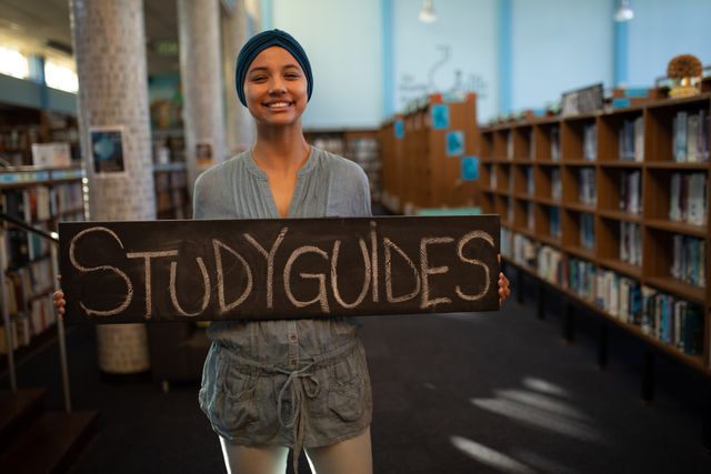 Front view of a biracial female student wearing a dark blue hijab studying in a library, holding a sign board with words study guides.
