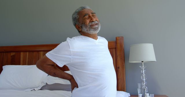 Elderly man clutching back in pain, sitting on bed in modern bedroom. Ideal for use in articles or advertisements about senior healthcare, pain relief, medical issues affecting the elderly, and home healthcare solutions. Can be used in contexts related to physical therapy, injury recovery, and promoting products designed for senior citizens' well-being.