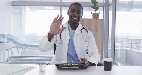 Smiling doctor waving while holding tablet and wearing stethoscope at bright modern clinic desk