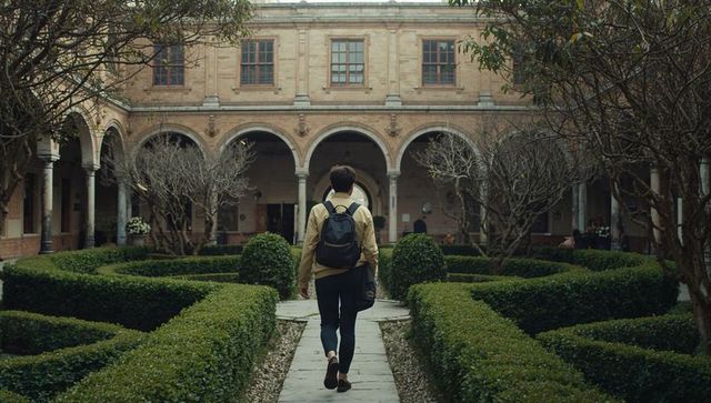 Young man walking cloister courtyard carrying backpack and shoulder bag, hedge symmetry