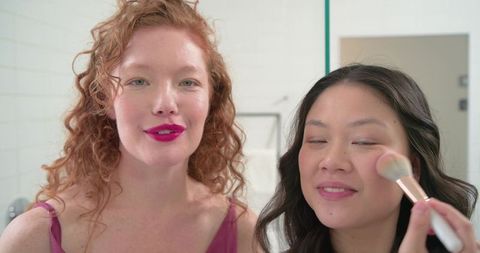 Redhead and asian friends applying makeup together in bright bathroom mirror