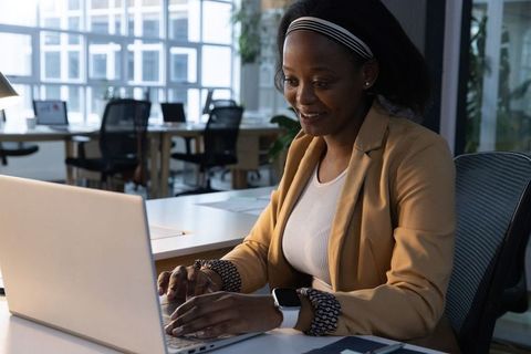 African american woman working on laptop in modern glass office with smartwatch