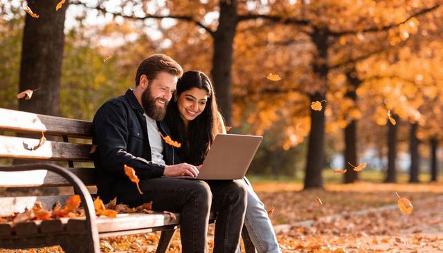 Couple browsing laptop on park bench in autumn with falling leaves