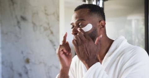 Mature man applying under-eye patch at bathroom vanity in white robe for skincare routine