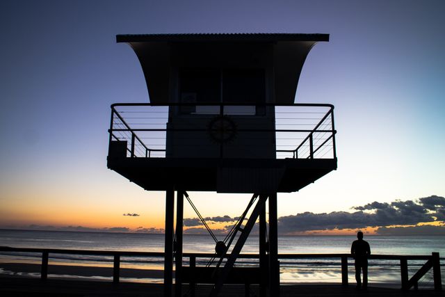 Perfect for travel websites, vacation advertisements, inspirational posters, and websites focused on beach activities or relaxation. The serene sunset combined with the lifeguard tower and silhouette of a person suggests tranquility and the ending of a peaceful day at the beach.