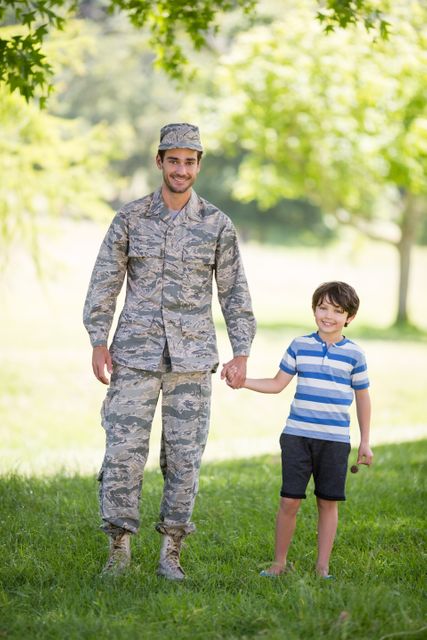 Army soldier in camouflage uniform holding hands with his young son in a park on a sunny day. Both are smiling and enjoying the outdoors. Ideal for use in family, military, and outdoor lifestyle themes, as well as promotional materials for military family support programs.