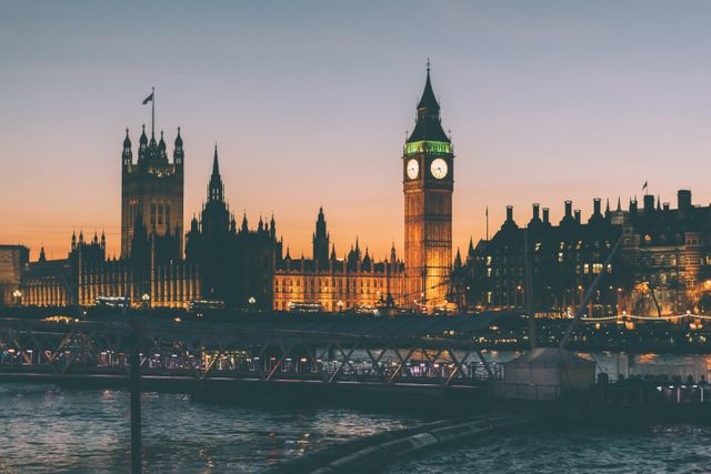View of Westminster Palace and Big Ben during sunset in London. The River Thames is in the foreground with illuminated buildings and reflected lights creating a serene evening atmosphere. Ideal for travel blogs, articles on London, historical features, and tourism promotion.