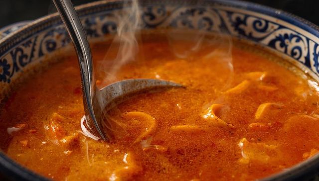 Steaming spicy noodle soup in blue patterned ceramic bowl with ladle and glossy broth
