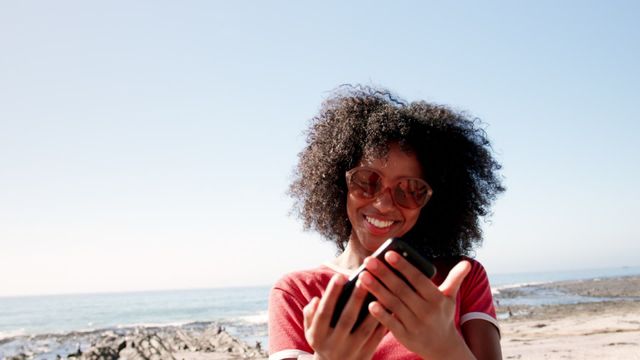 Young African descent woman poses for a selfie while relaxing on the beach. Captured on a sunny day, her cheerful smile and the tranquil ocean in the backdrop exude the essence of a perfect vacation. This image is suitable for travel and leisure brochures, mobile phone promotions, or social media content highlighting connectivity and joyful moments.