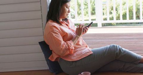 Mature asian woman relaxing on sunlit porch using smartphone for messaging and browsing