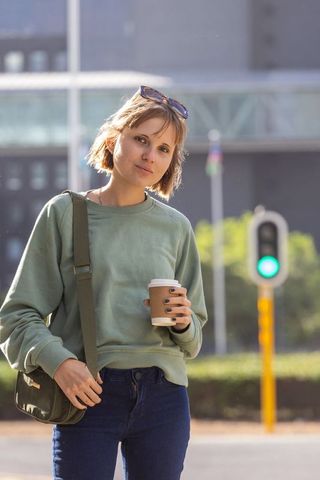 Young woman standing on urban sidewalk holding takeaway coffee wearing green sweatshirt