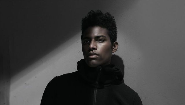 Moody low-key portrait of male-presenting model standing in diagonal spotlight and shadow