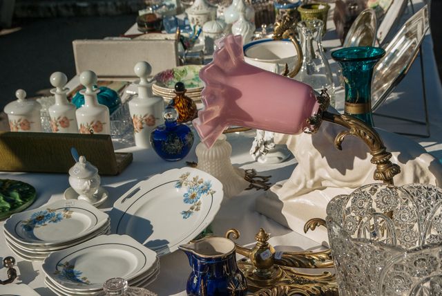 Antique porcelain and glassware displayed on tables at an outdoor flea market, a mix of plates, lamps, and other decorative items. Ideal for use in articles or advertisements related to antique collecting, flea markets, and vintage home decor. Suitable for blogs or websites focused on shopping, home decor, and antiques.