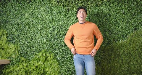 Young asian man standing against lush vertical garden wearing orange sweatshirt and jeans