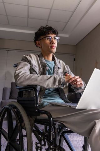 Young asian man sitting in wheelchair using laptop in home workspace, inclusive tech