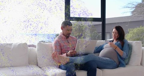 Expectant couple relaxing on sunlit sofa, partner browsing laptop while expectant woman sipping mug