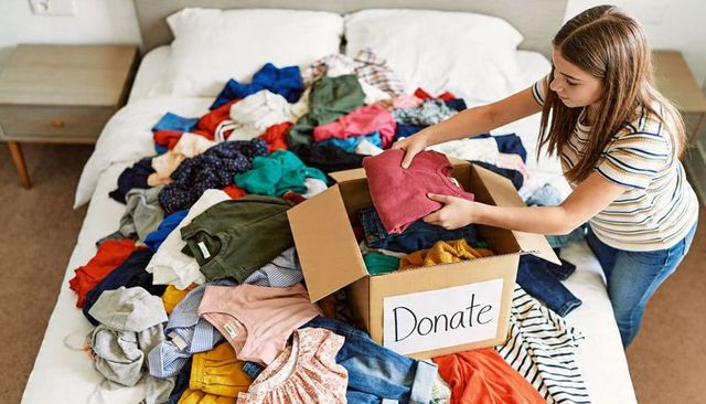 Teen girl sorting clothes for donation on bed packing box for charity and decluttering